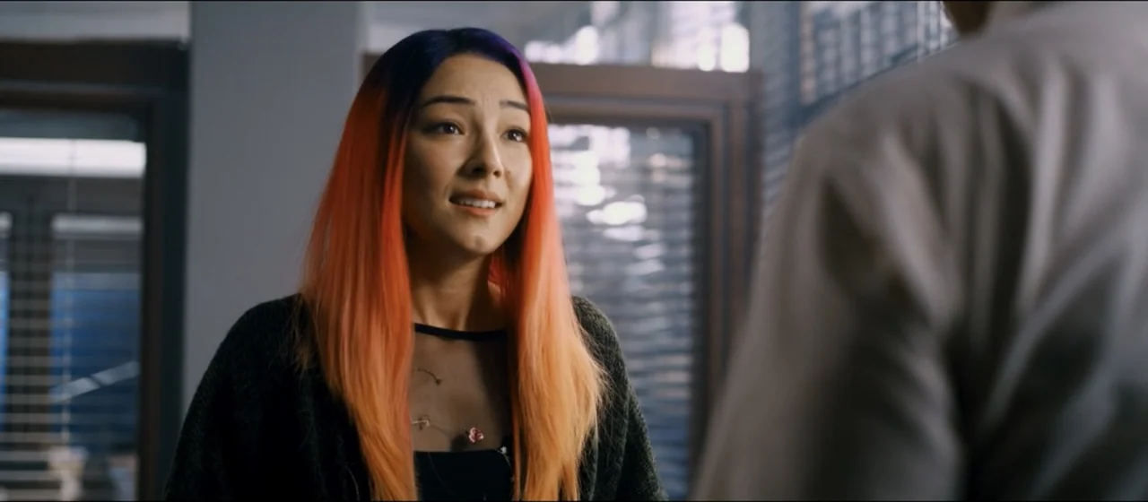 Guns Akimbo (2020) movie still: medium — An interior office space with blurred window blinds in the background. A young woman with colorful hair…