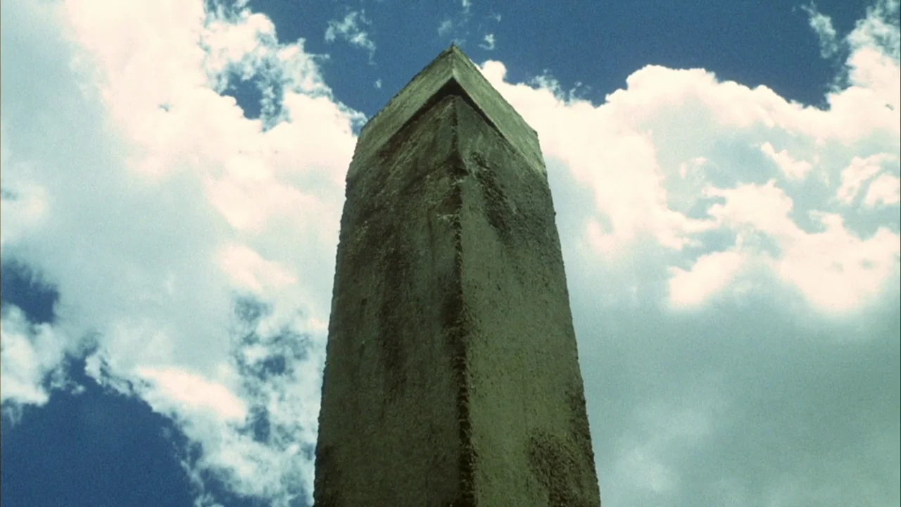 Phase IV (1974) movie still: wide — A tall, rectangular concrete obelisk stands isolated under a blue, cloud-filled sky. The weathered,…