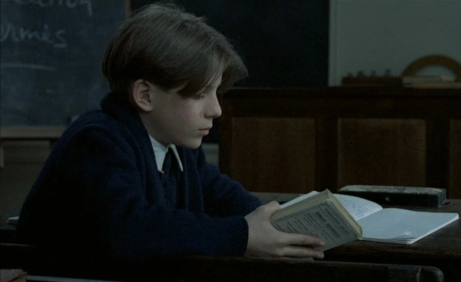 Au Revoir Les Enfants (1987) movie still: medium — A dimly lit classroom features old wooden desks and a dark blackboard in back. A young boy in a…