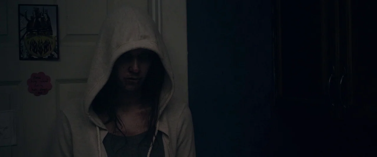 Starry Eyes (2014) movie still: medium — A dimly lit interior space features a white door with a small framed photo attached. A person wearing a…