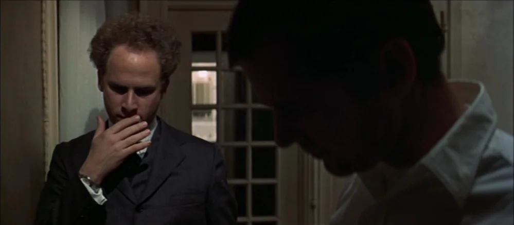 Carnal Knowledge (1971) movie still: medium — A dimly lit narrow hallway with a glass-paned door visible in the background. Two men stand facing…