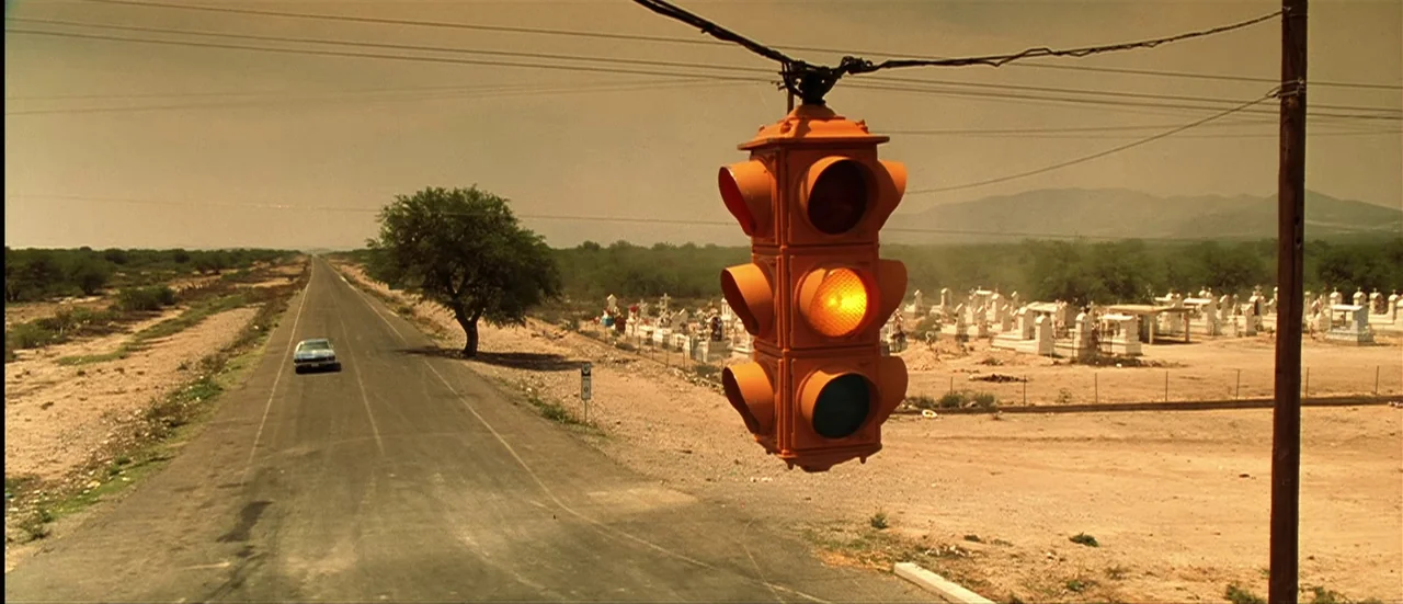 The Mexican (2001) movie still: establishing — A long, flat asphalt road stretches into a sparse, arid desert landscape at midday. A yellow traffic…