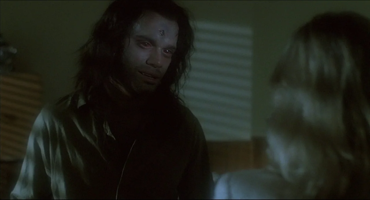 The Howling (1981) movie still: medium — A dark indoor space with striped shadows cast by window blinds on the wall. A man with a metallic mark on…