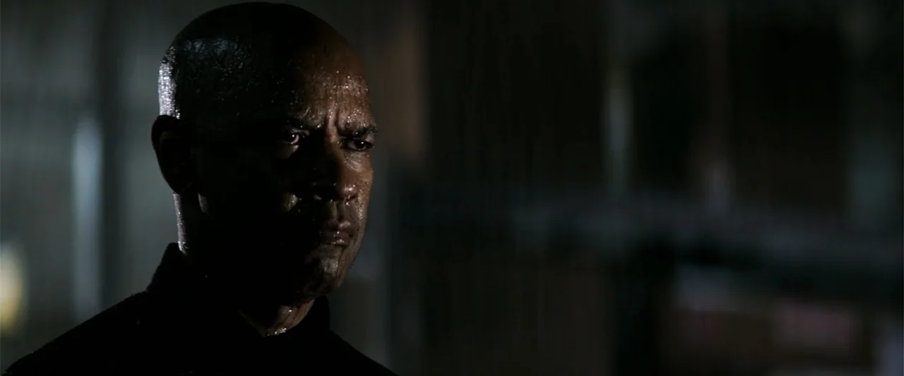 The Equalizer (2014) movie still: closeup — A man stands in a dark outdoor setting at night while it rains. The man looks toward the side of the…