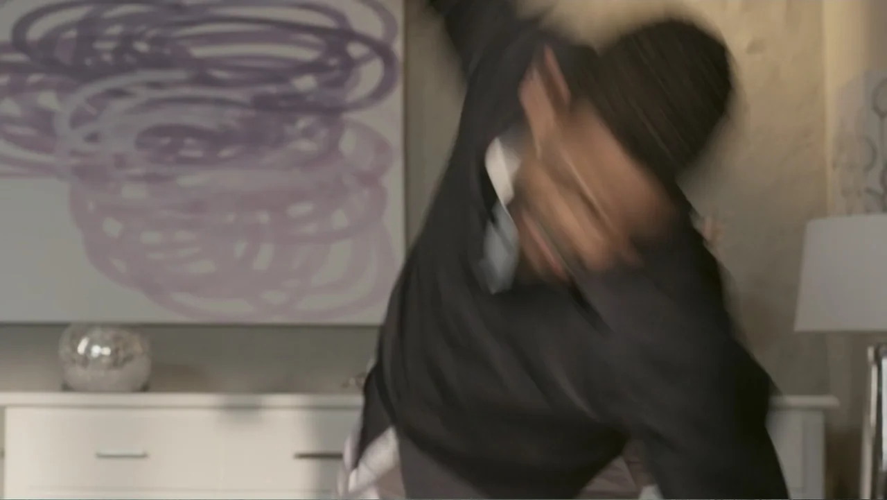 Locas y atrapadas (2014) movie still: medium — A modern living room features a white sideboard and an abstract purple wall art piece. A man moves…