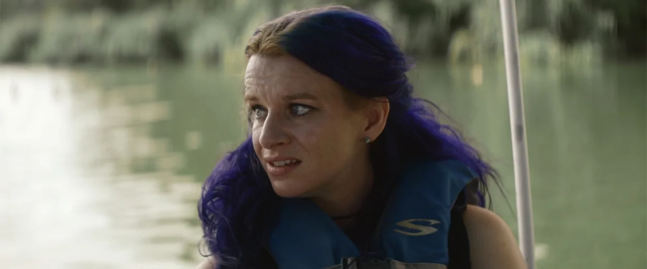 Mennonite's Bite (2023) movie still: medium — A person is on a boat on open water during the day. The woman with purple hair wearing a blue life…