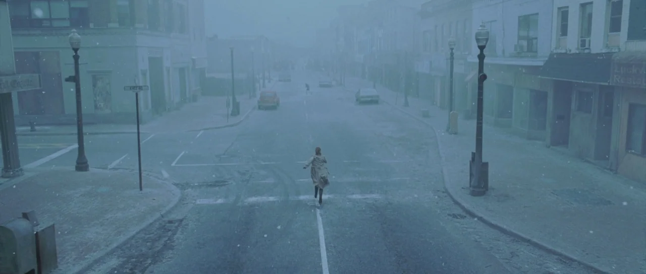 Silent Hill movie still: wide — An empty intersection in a desolate small town is covered in light snowfall. A lone person in a trench coat runs…