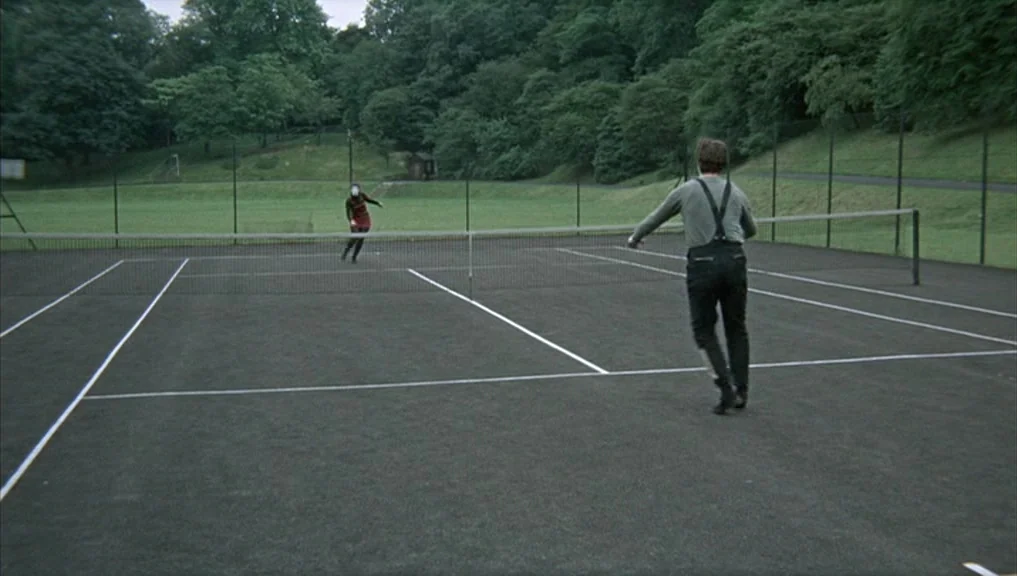 Blow-Up (1966) movie still: wide — A dark-surfaced tennis court surrounded by a chain-link fence and dense green trees. Two individuals engage in a…