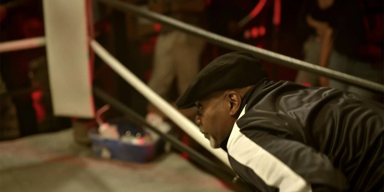 7th & Union (2021) movie still: medium — A man in a track jacket leans over the ropes of a ring. He looks towards the center of the ring in a dark…