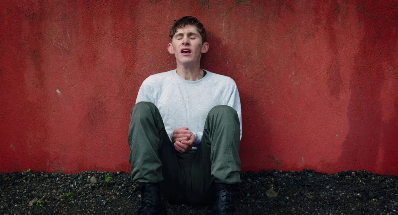 Dating Amber movie still: fullbody — A man sits against a textured red wall on a dark gravel patch. He has his eyes closed and his hands are held…