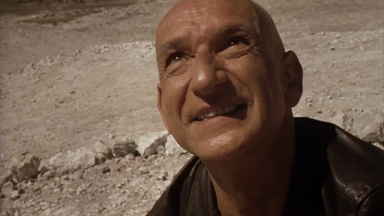Suspect Zero (2004) movie still: closeup — A dry desert landscape with rocky ground under a bright daytime sky. An older man gazes upward at…