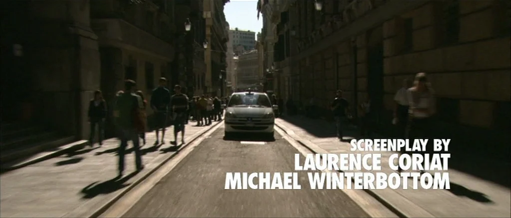 Genova (2008) movie still: wide — A narrow European-style urban street lined with multi-story stone buildings and dark awnings. A white car…