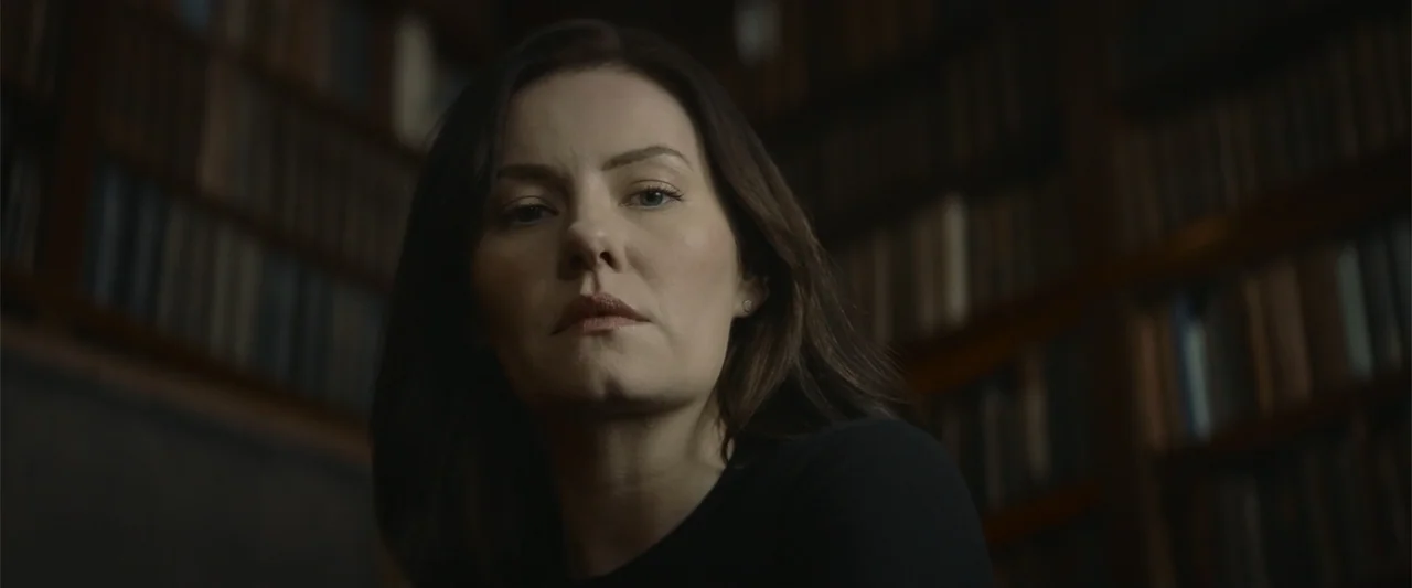 The Cellar (2022) movie still: closeup — A dark, dimly lit library with rows of blurry books lining the background shelves. A woman with dark hair…
