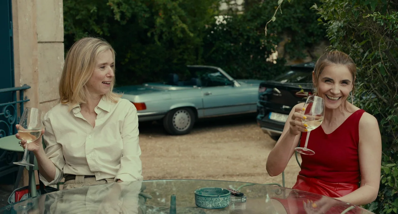 Last Summer (2023) movie still: medium — Two women sit at a glass patio table outdoors with wine glasses in hand. Behind them a light blue vintage…