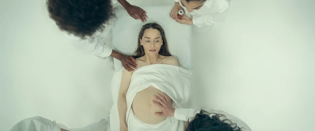 The Pod Generation (2023) movie still: wide — A stark white, brightly lit clinical room contains a patient lying on a table. Four people in white…