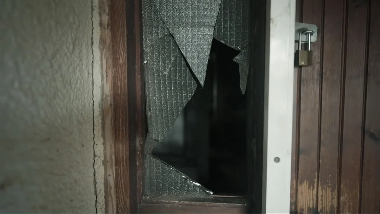 The Children of October 7 (2025) movie still: closeup — A wooden door features a panel of textured glass shattered into large triangular fragments.…
