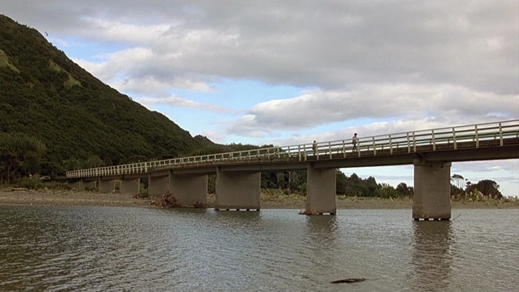 Boy (2010) movie still: wide — A long concrete bridge crosses a wide river with a green, forested hill nearby. The sky is filled with clouds over…