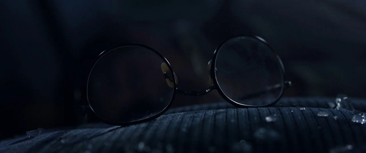 Shazam! (2019) movie still: closeup — A pair of round glasses sits on a dark ribbed seat fabric in darkness. Small shards of broken glass are…