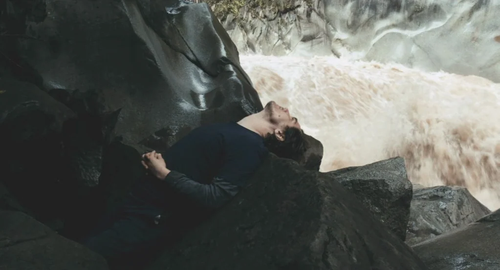 My Son, My Son, What Have Ye Done? (2009) movie still: medium — A man rests against a smooth boulder at the edge of a turbulent river. Rapid white…