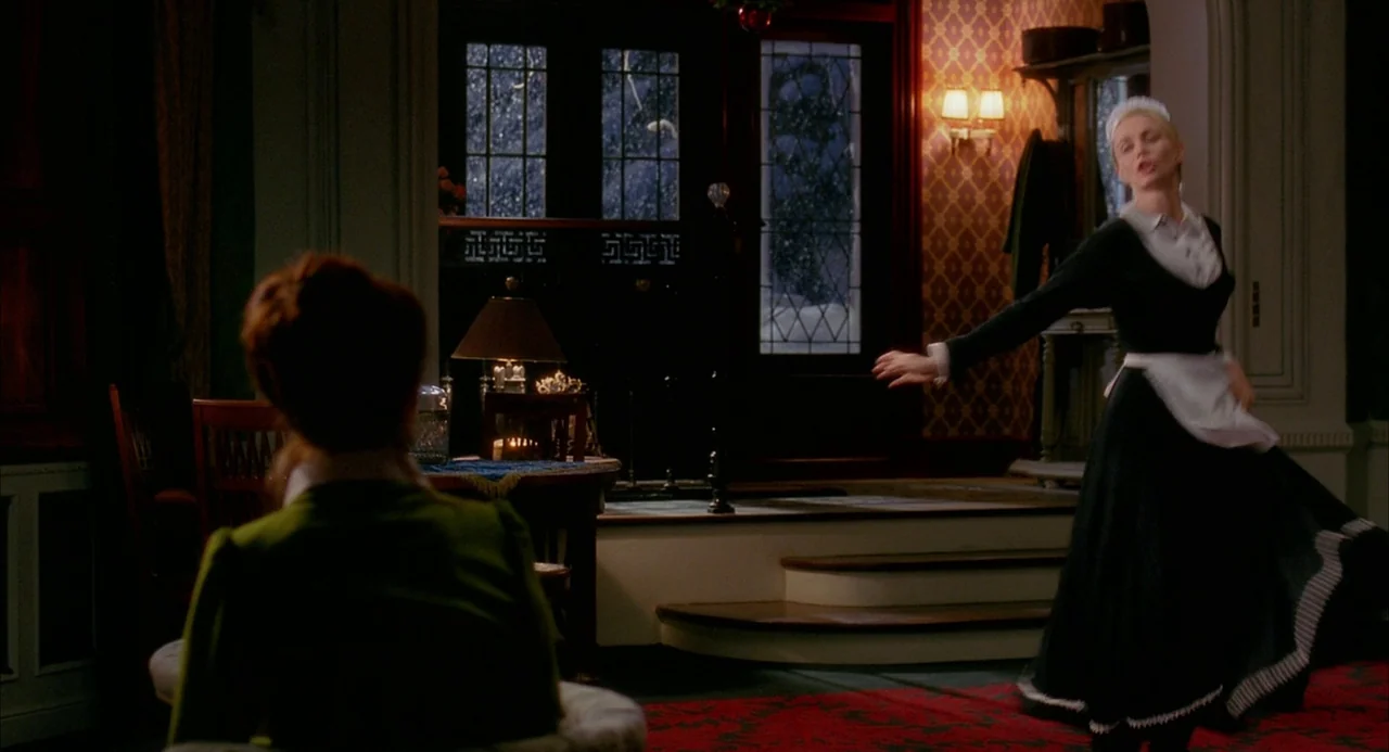 8 Women (2002) movie still: medium — A dark, elegant entry hall with wallpaper, wooden steps, and a glass-paneled front door. A maid stands in the…