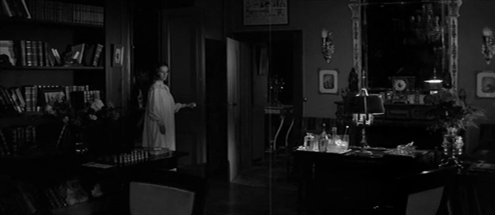 Les Amants (1958) movie still: wide — A dark, wood-paneled room is filled with bookshelves and a desk with a lamp. A woman wearing a nightgown…