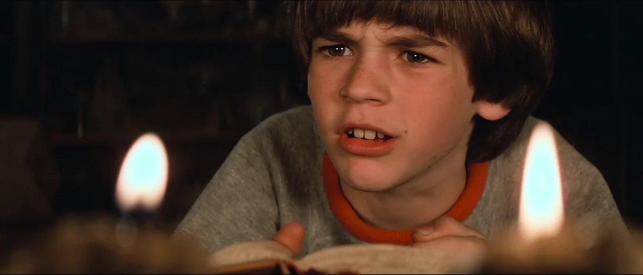 The NeverEnding Story (1984) movie still: closeup — A dimly lit room with a young boy looking down at an open book. The scene is illuminated by two…