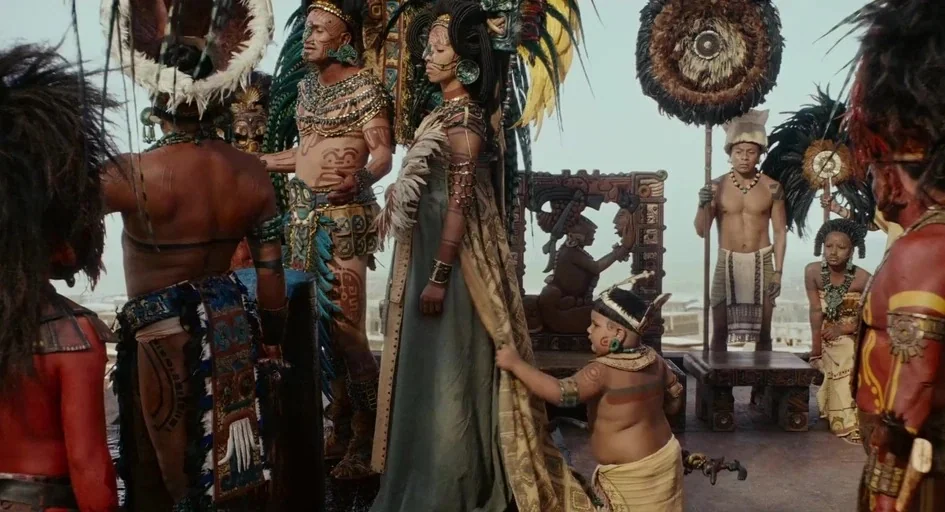 Apocalypto (2006) movie still: wide — A stone courtyard elevated above a city landscape holds several figures in ceremonial garb. A queen stands…
