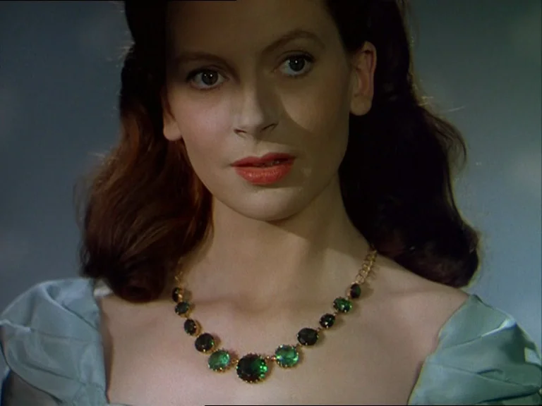 Black Narcissus (1947) movie still: closeup — A close-up portrait shows a woman in a light blue dress with an emerald necklace. The background is a…