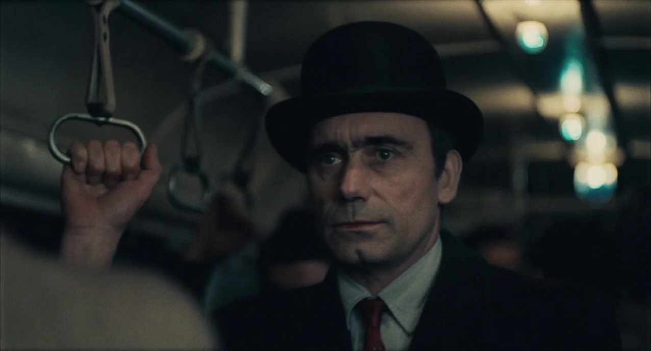 The Army of Shadows (1969) movie still: closeup — A dimly lit subway car interior with rows of hanging hand straps. A middle-aged man stands in the…