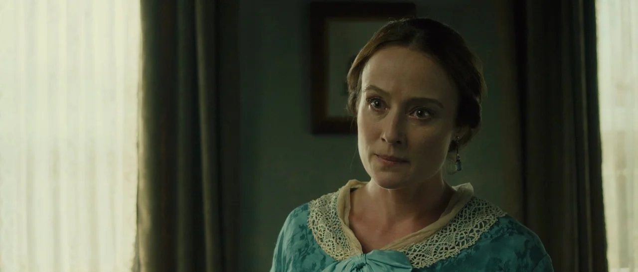 A Quiet Passion (2016) movie still: medium — A dimly lit room with muted green walls and a framed print on the wall. A woman in a blue…