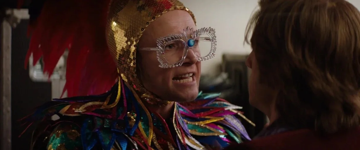 Rocketman (2019) movie still: medium — A cramped dressing room filled with indistinct stage equipment and dim, practical light. A man wearing a…