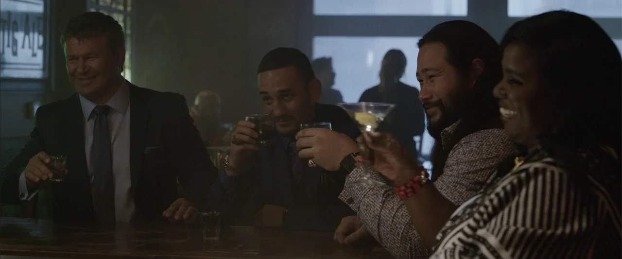 Den of Thieves (2018) movie still: medium — A dimly lit bar setting with people gathered around a wooden counter. Four individuals sit together…