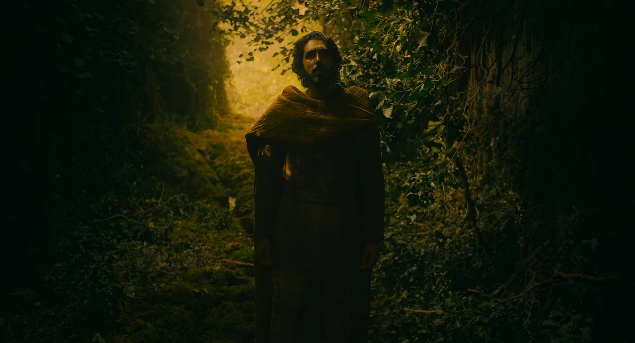 The Green Knight (2021) movie still: medium — A dark, narrow path is densely overgrown with moss and thick green leaves. A man with a beard stands…