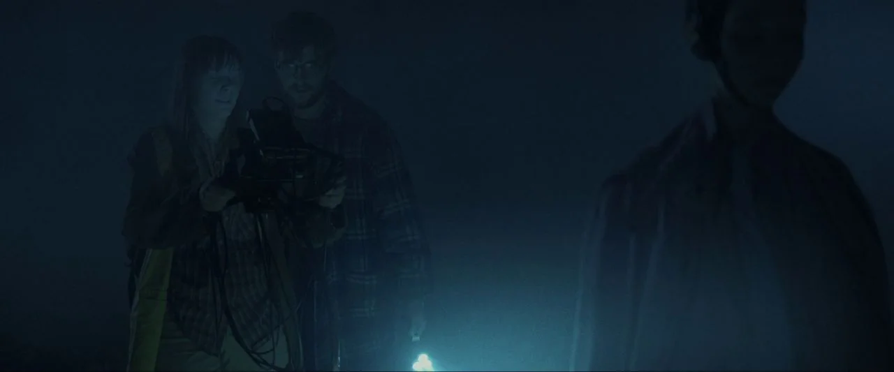 Come True (2020) movie still: medium — A dark, foggy outdoor field is illuminated by the faint glow of technology. Two people hold electronic…