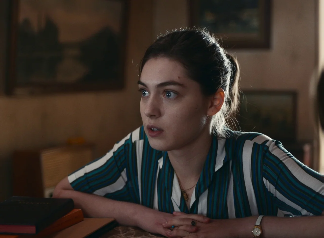 Happening (2021) movie still: medium — An indoor space features framed wall art and warm-toned surfaces in the background. A woman leans over a…