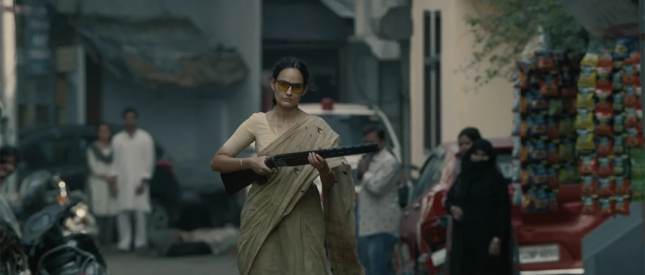 Nishaanchi 2 (2025) movie still: medium — A busy urban street features parked vehicles and pedestrians in the blurred background. A woman dressed in…