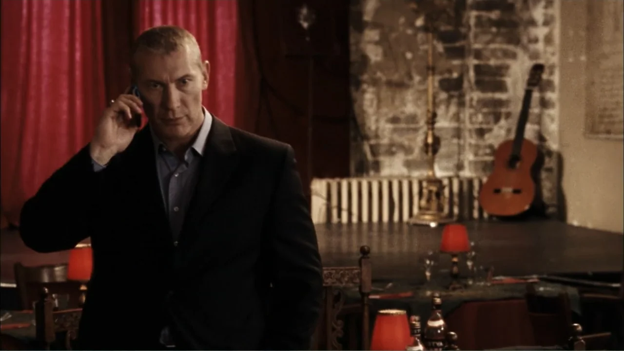 Driven to Kill (2009) movie still: medium — A dimly lit restaurant interior features brick walls and a small wooden stage area. A middle-aged man in…