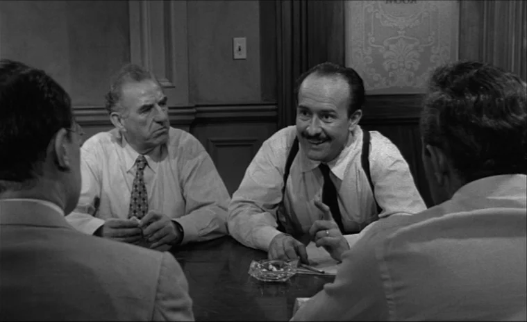 12 Angry Men (1957) movie still: A tense deliberation scene featuring men at a table, framed by foreground figures to establish a claustrophobic…