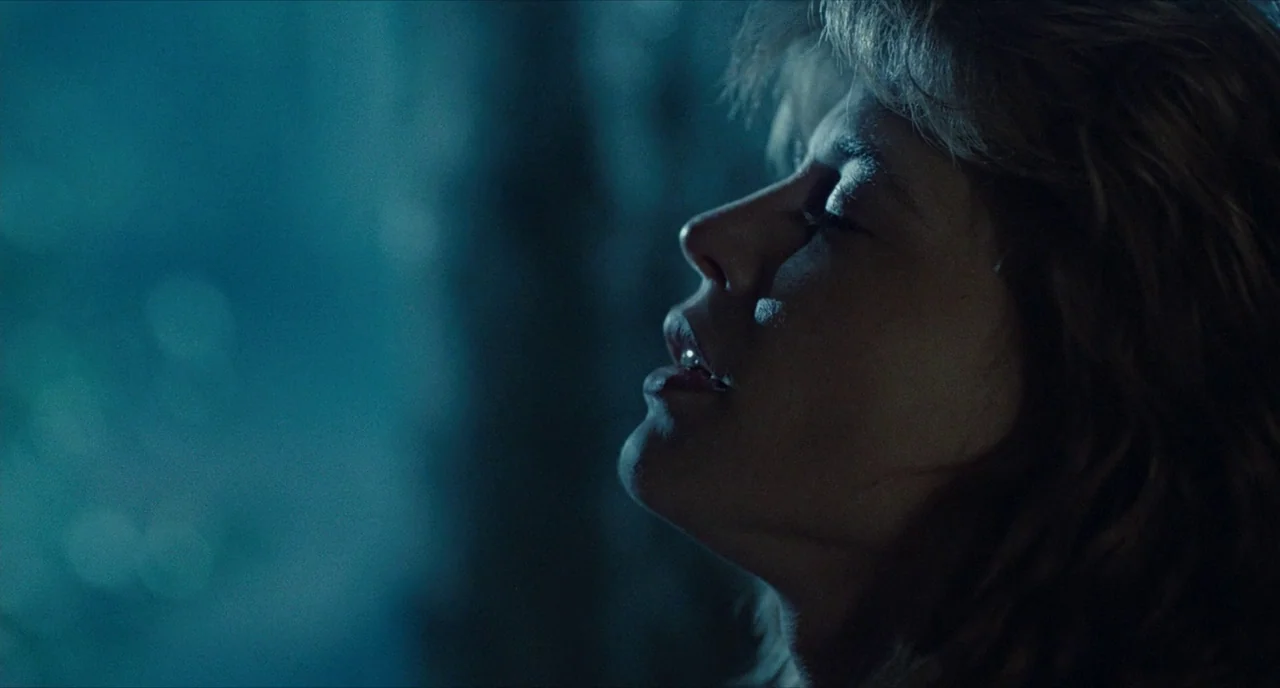 The Terminator (1984) movie still: closeup — The face of a woman is visible in darkness against a blurry blue background. She tilts her head upward…