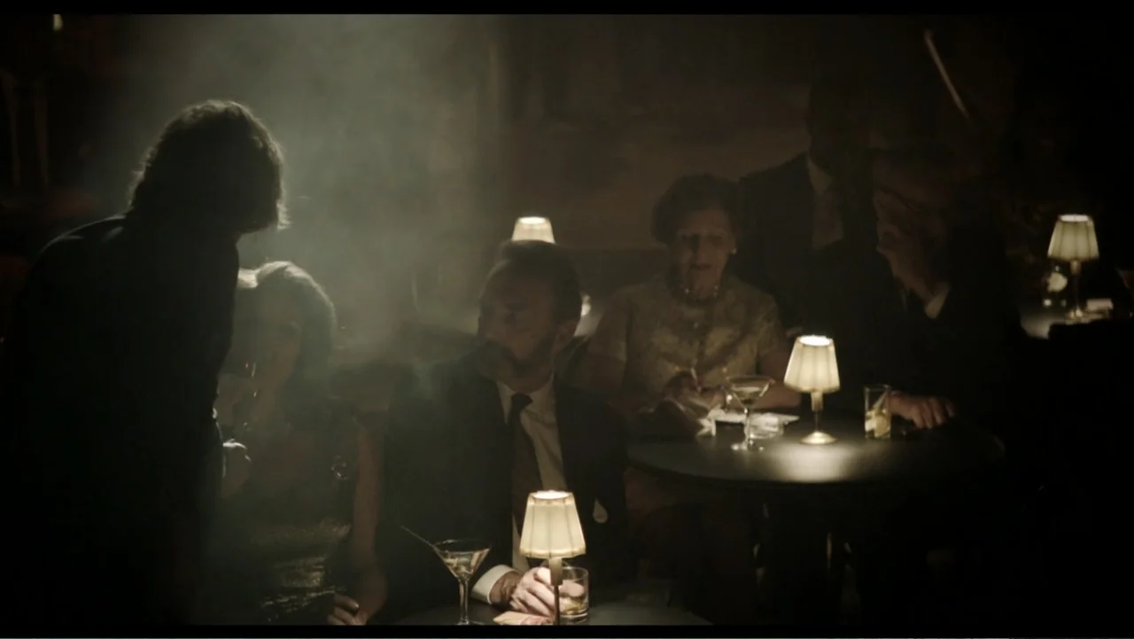 Six by Sondheim (2013) movie still: medium — A dimly lit club features small round tables each with a glowing lamp on top. Several people sit at…