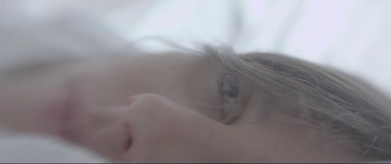 Sun Choke (2015) movie still: closeup — A close-up view of a person lying down on white bedding. Only a portion of the face is visible in the frame.