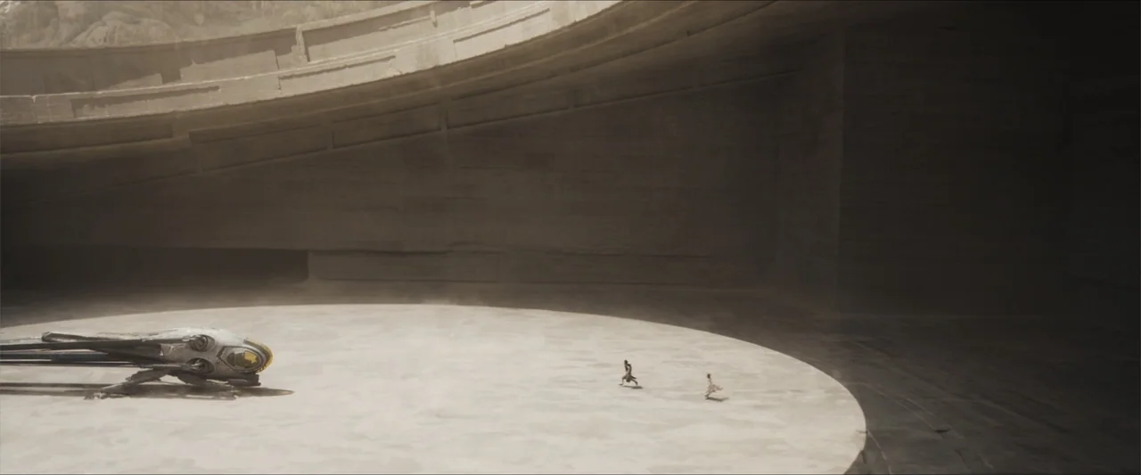 Dune (2021) movie still: wide — A vast, curved concrete structure encloses a circular landing pad in a desert. Two figures run across the flat…