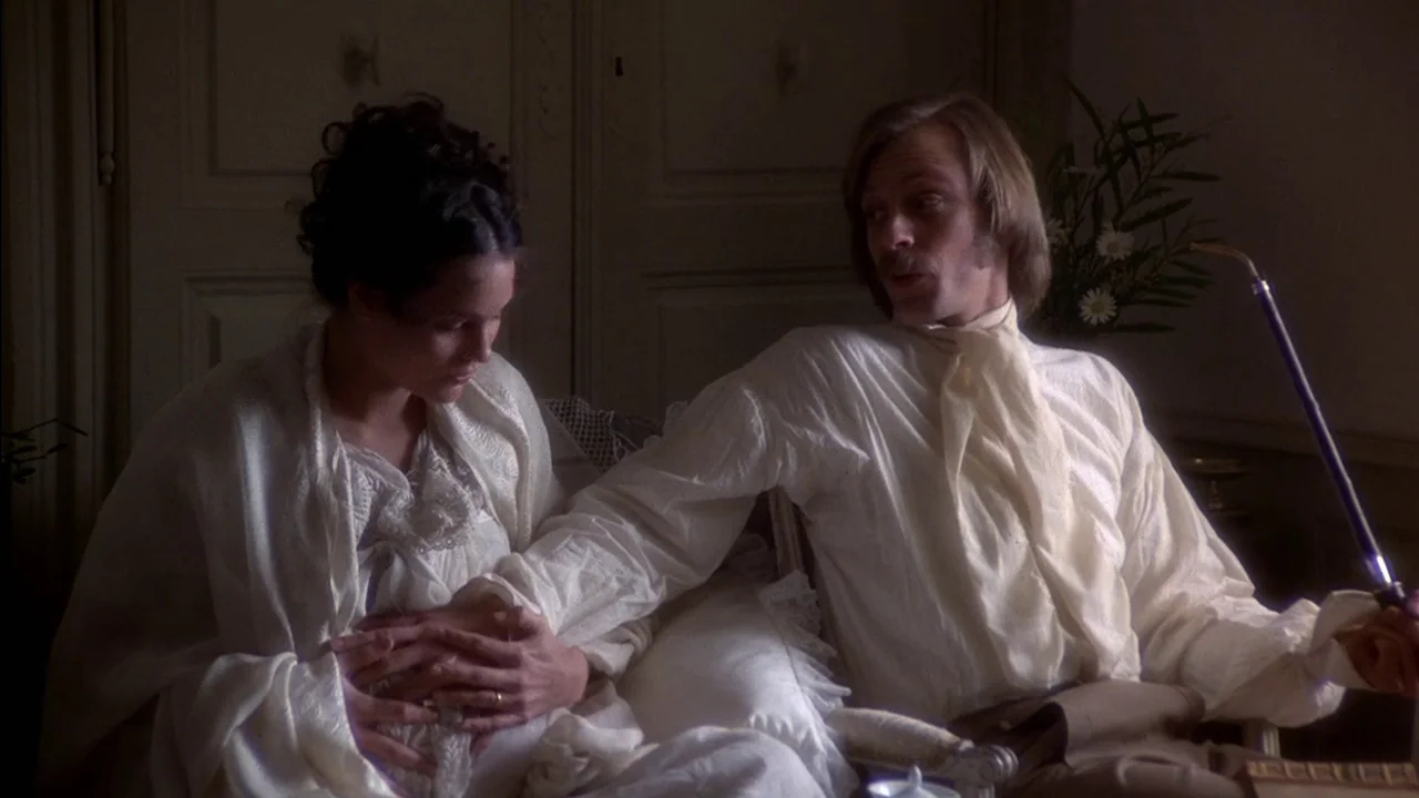 The Duellists (1977) movie still: medium — A dimly lit room with light-colored wooden doors in the background. A man sits next to a woman and holds…