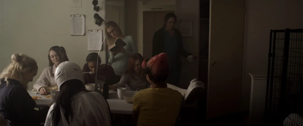 Mermaid Down (2019) movie still: medium — A dimly lit room with several women sitting at a folding plastic table. Several individuals are eating…