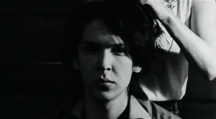 Roadkill (1989) movie still: closeup — A dark indoor space with high-contrast lighting obscuring the background. A person reaches into the frame to…