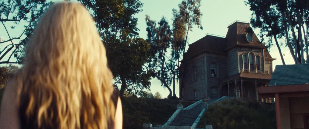 MaXXXine (2024) movie still: medium — A Victorian-style wooden mansion sits atop a grassy hill with concrete steps leading up. A blonde woman in the…