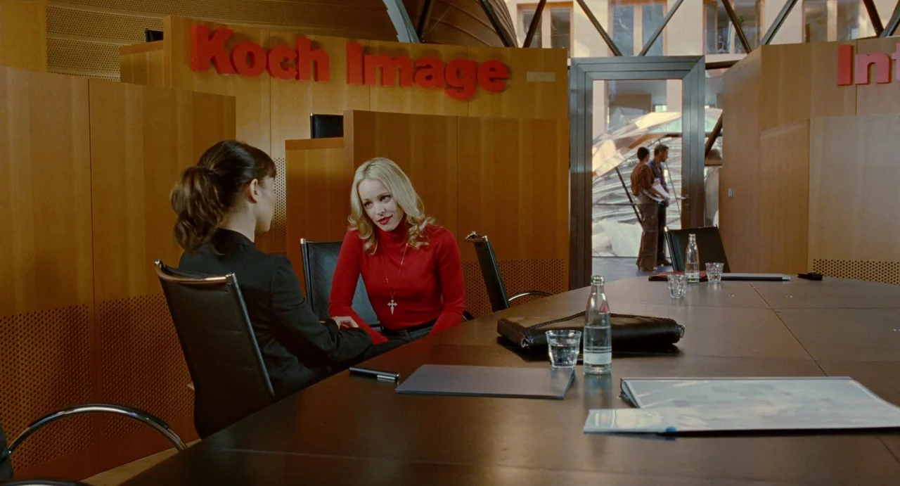 Passion (2012) movie still: medium — A modern corporate office features light wood paneling and a large dark conference table. Two young women sit…