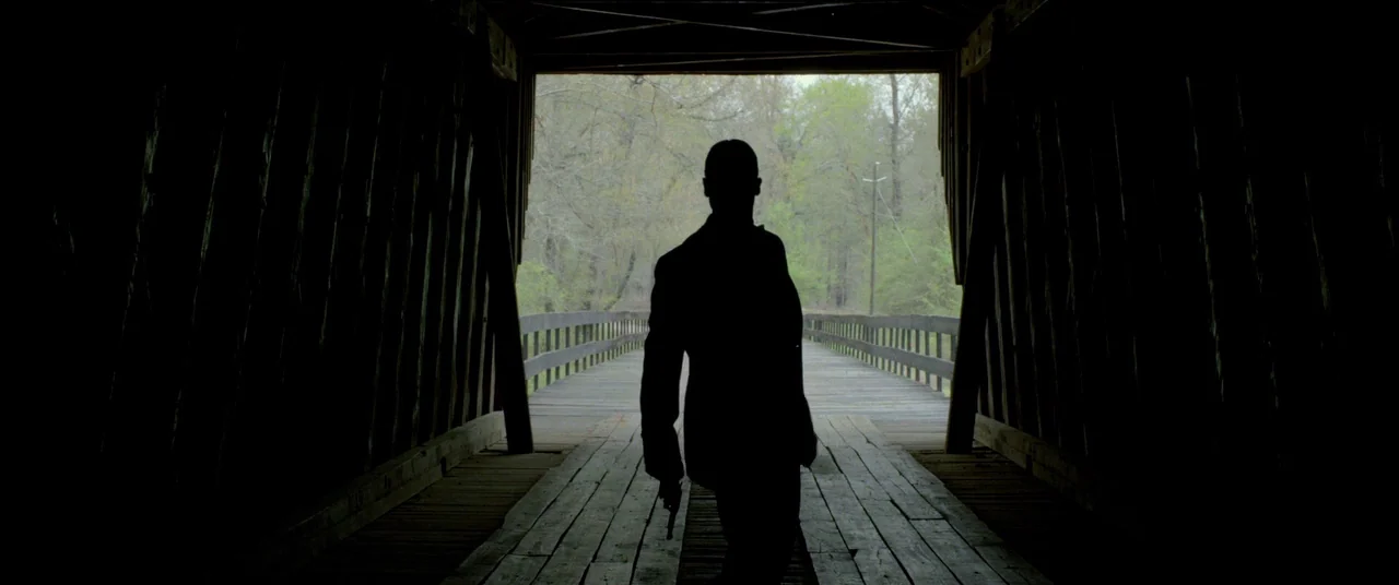 Lawless (2012) movie still: fullbody — A long wooden covered bridge leads to an open outdoor area with trees visible. A man in a suit stands in the…