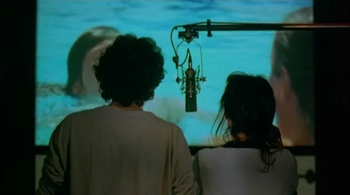 Code Unknown (2000) movie still: medium — A dark recording booth features a large projection screen displaying a swimming scene. Two people stand…