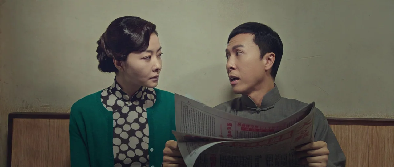 Ip Man 3 (2015) movie still: medium — A man and woman sit side by side against a plain, off-white wall. The man holds an open newspaper and speaks…